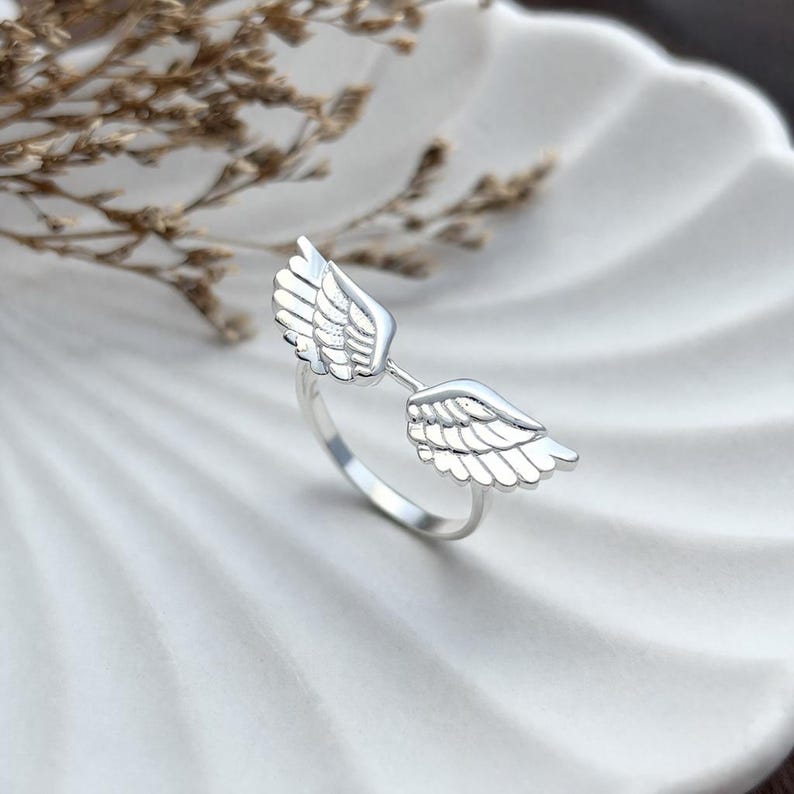 May include: A silver ring featuring a pair of detailed angel wings. The wings are positioned on either side of the ring's band, creating an open design. The ring is displayed on a white, textured surface.