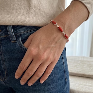Può includere: Un bracciale in argento con cinque pietre rettangolari di corallo rosso vivo. Il bracciale è indossato su un polso, con la mano in una tasca di jeans blu. Lo sfondo è beige e bianco neutro.