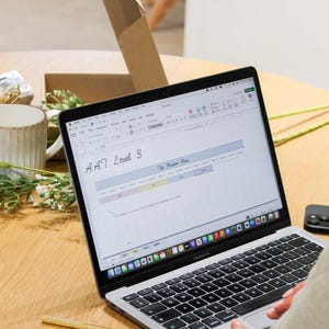 May include: A silver MacBook Air laptop displaying a spreadsheet titled "A47 Level 3" and "The Master Plan". A gold pen and a black phone are on the wooden table. A person's hands are visible, along with a ring. A cardboard box and a mug are in the background.