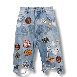 Op de afbeelding: Lichtblauwe spijkerbroek met versleten plekken, gerafelde zomen en meerdere geborduurde patches. De patches bevatten ronde en rechthoekige ontwerpen met de tekst SPBY, 1999 en diverse iconen. De letters SPBY zijn in een groot lettertype op de voorkant gestikt.