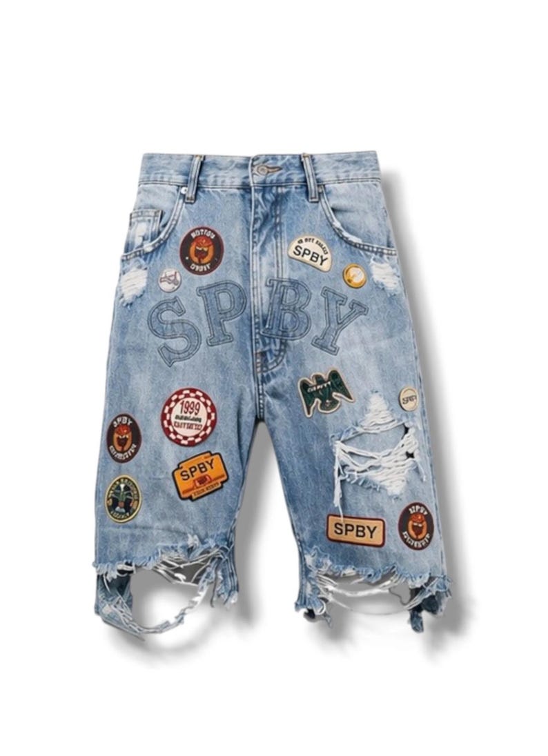 Op de afbeelding: Lichtblauwe spijkerbroek met versleten plekken, gerafelde zomen en meerdere geborduurde patches. De patches bevatten ronde en rechthoekige ontwerpen met de tekst SPBY, 1999 en diverse iconen. De letters SPBY zijn in een groot lettertype op de voorkant gestikt.