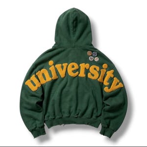 Op de afbeelding: Een donkergroene hoodie met het woord university in grote, textuurrijke gele letters op de achterkant. Het kledingstuk heeft een capuchon, lange mouwen en vier kleine ronde pins bevestigd aan de rechterbovenkant van de achterzijde.