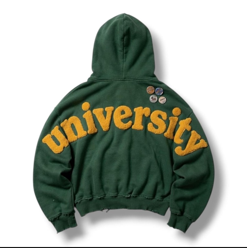 Op de afbeelding: Een donkergroene hoodie met het woord university in grote, textuurrijke gele letters op de achterkant. Het kledingstuk heeft een capuchon, lange mouwen en vier kleine ronde pins bevestigd aan de rechterbovenkant van de achterzijde.
