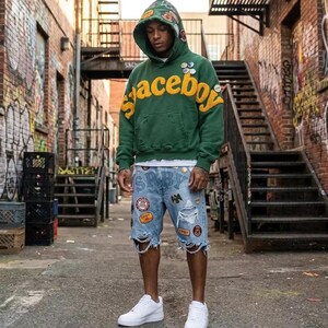 Op de afbeelding: Een groene hoodie met de gele tekst spaceboy op de borst, voorzien van diverse patches en een gerafelde zoom. Gecombineerd met een lichtblauwe spijkerbroek met een versleten afwerking, meerdere patches en witte sneakers.