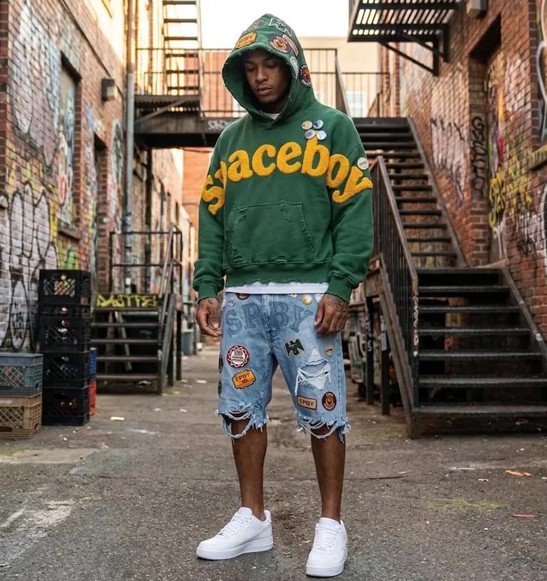 Op de afbeelding: Een groene hoodie met de gele tekst spaceboy op de borst, voorzien van diverse patches en een gerafelde zoom. Gecombineerd met een lichtblauwe spijkerbroek met een versleten afwerking, meerdere patches en witte sneakers.