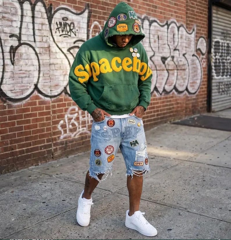 Op de afbeelding: Een groene hoodie met gele tekst spaceboy en diverse patches op de capuchon, gecombineerd met lichtblauwe versleten denim shorts met meerdere kleurrijke patches, geborduurde tekst SPBY en witte sneakers met een laag model.