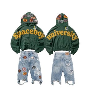Op de afbeelding: Een donkergroene hoodie met gele geborduurde tekst Spaceboy University en diverse kleurrijke patches op de capuchon, gecombineerd met een lichtblauwe distressed denim short met meerdere patches en gerafelde zomen.