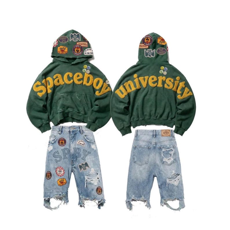 Op de afbeelding: Een donkergroene hoodie met gele geborduurde tekst Spaceboy University en diverse kleurrijke patches op de capuchon, gecombineerd met een lichtblauwe distressed denim short met meerdere patches en gerafelde zomen.