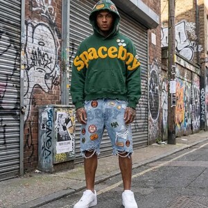 Op de afbeelding: Een donkergroene hoodie met het woord spaceboy in grote gele letters op de borst, voorzien van diverse kleine ronde pins en versleten details. Gecombineerd met een lichtblauwe spijkerbroek met verschillende patches en scheuren.
