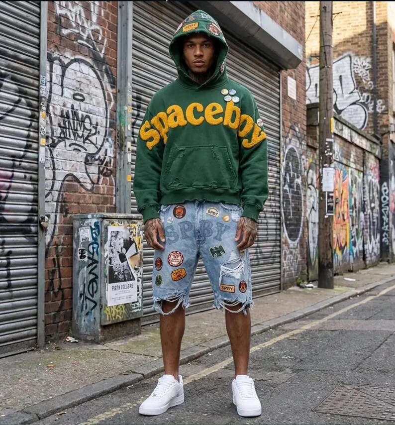 Op de afbeelding: Een donkergroene hoodie met het woord spaceboy in grote gele letters op de borst, voorzien van diverse kleine ronde pins en versleten details. Gecombineerd met een lichtblauwe spijkerbroek met verschillende patches en scheuren.