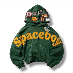Op de afbeelding: Een donkergroene hoodie met het woord Spaceboy in grote, textuurrijke gele letters op de borst. De capuchon is versierd met meerdere kleurrijke geborduurde patches en het kledingstuk heeft versleten randen en kleine ronde speldjes.