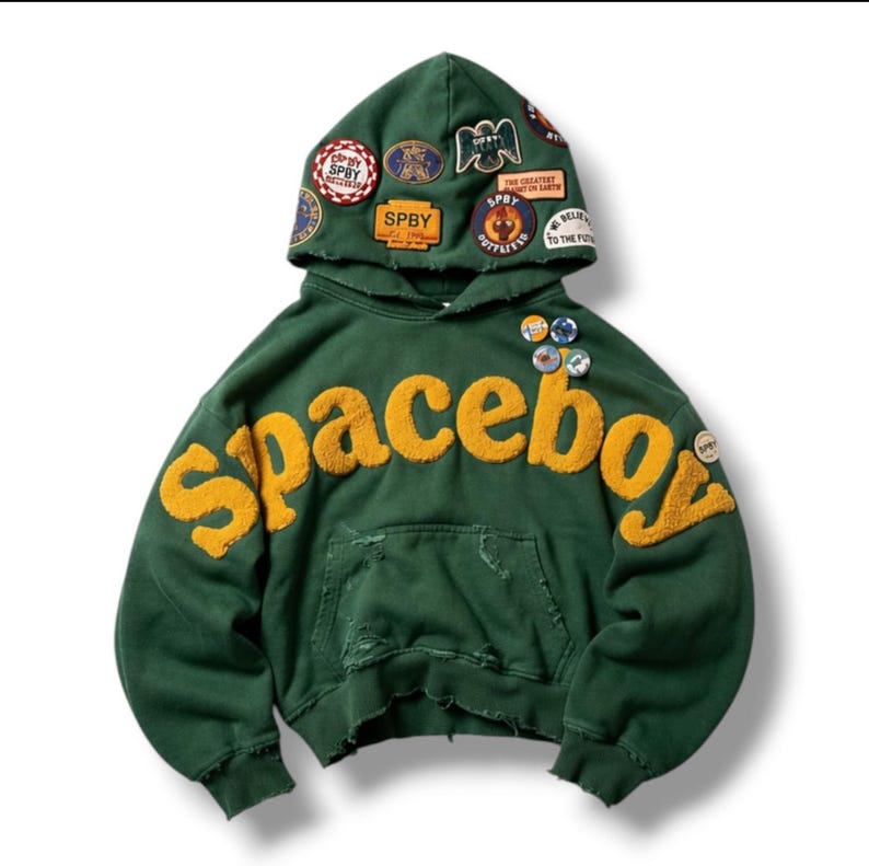 Op de afbeelding: Een donkergroene hoodie met het woord Spaceboy in grote, textuurrijke gele letters op de borst. De capuchon is versierd met meerdere kleurrijke geborduurde patches en het kledingstuk heeft versleten randen en kleine ronde speldjes.