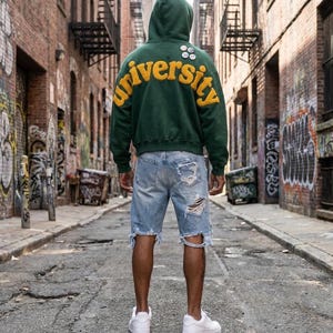 Op de afbeelding: Een donkergroene hoodie met het woord university in grote gele vilten letters op de achterkant, gecombineerd met lichtblauwe distressed denim shorts met gerafelde zomen en scheuren, en strakke witte lage sportschoenen.