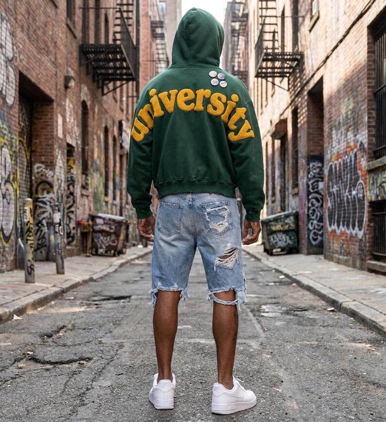 Op de afbeelding: Een donkergroene hoodie met het woord university in grote gele vilten letters op de achterkant, gecombineerd met lichtblauwe distressed denim shorts met gerafelde zomen en scheuren, en strakke witte lage sportschoenen.