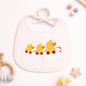 May include: White baby bib with a quilted texture and a tie closure. The bib features an embroidered design of three yellow ducklings on red wheels. The ducks are connected by a string with green and red beads. A pacifier and wooden star are also visible.
