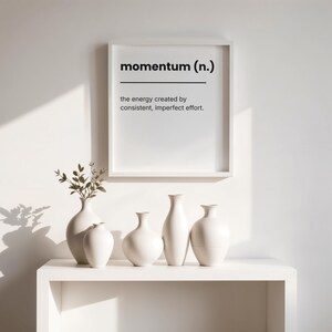 Puede incluir: Una impresión enmarcada en blanco con la palabra "momentum" y su definición cuelga sobre una estantería blanca. Cinco jarrones de cerámica blanca de diferentes formas y tamaños están colocados en la estantería. Un jarrón con vegetación está a la izquierda.