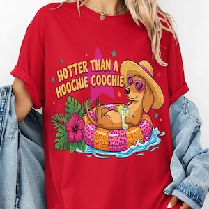 Può includere: Maglietta rossa con una grafica di un bassotto con occhiali da sole e un cappello di paglia, che si rilassa su un anello gonfiabile in acqua. Il testo "HOTTER THAN A HOOCHIE COOCHIE" è sopra il cane.