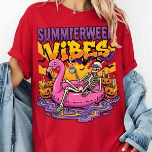 Peut inclure: T-shirt rouge avec un motif graphique représentant un squelette se relaxant sur une bouée flamant rose, tenant une tasse, avec le texte "Summerween Vibes". Le motif comprend des chauves-souris, des citrouilles et des bougies.
