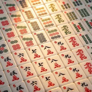 May include: A collection of off-white Mahjong tiles arranged in a grid, featuring various red, green, and black engraved symbols, including Chinese characters, numerical digits, floral patterns, and dragon illustrations on the face of each tile.