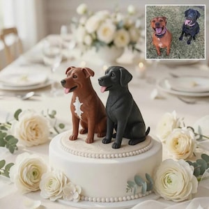 Könnte beinhalten: Eine weiße Hochzeitstorte mit zwei Hundefiguren, eine braune und eine schwarze. Die braune Figur hat eine weiße Markierung auf der Brust. Die Torte ist mit weißen Blumen und Grünzeug dekoriert. Ein Foto der Hunde befindet sich oben rechts.