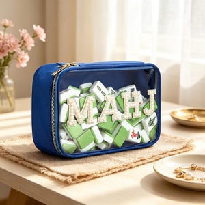 May include: A rectangular blue storage pouch with a clear front panel containing a set of green and white mahjong tiles. The front of the pouch features the letters MAHJ in large, white, pearl-embellished patches attached to the clear surface.