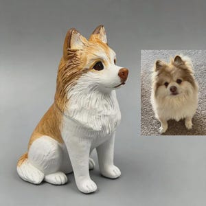 Puede incluir: Una figura de perro de cerámica en posición sentada, con una combinación de colores blanco y marrón. El perro tiene una nariz marrón y ojos oscuros. Una foto más pequeña de un perro real está en el fondo.