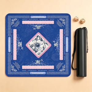Puede incluir: Una alfombrilla de juego cuadrada azul con un diamante central de patrón floral en blanco y negro, rodeado de pistas de puntuación rectangulares rosas, direcciones de brújula y bordes decorativos. Incluye dos dados de madera y un estuche negro.