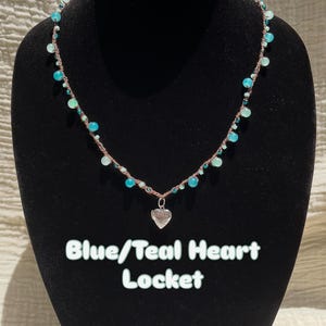May include: A necklace featuring a silver heart locket suspended from a brown cord adorned with various blue and teal beads. The text Blue/Teal Heart Locket is displayed below the locket. The design incorporates small round beads in shades of turquoise and aqua, creating a colorful contrast against the neutral cord.