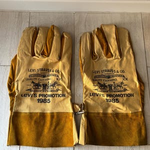 May include: A pair of tan and brown leather work gloves. The gloves have the Levi Strauss & Co. logo and "Levi's Promotion 1985" printed on them. The gloves are made of a durable material and have a metal clasp.