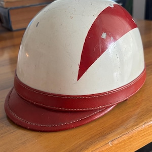 Pode incluir: Capacete branco vintage com design de faixa vermelha e viseira vermelha. O capacete mostra sinais de desgaste, indicando que é antigo. A viseira é feita de material vermelho semelhante ao couro. Este capacete é provavelmente para esportes ou atividades recreativas.
