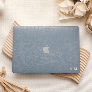 May include: A slate blue laptop case with a textured crocodile skin pattern and a silver Apple logo cutout in the center. The bottom right corner features the gold embossed initials K.M. The case has a rectangular shape with rounded corners.