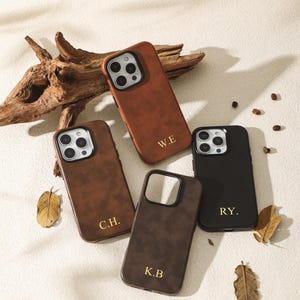 Pode incluir: Quatro capas de telefone de couro em tons de castanho e preto, cada uma com iniciais douradas. As capas estão dispostas sobre uma superfície neutra com folhas secas e um pedaço de madeira flutuante.