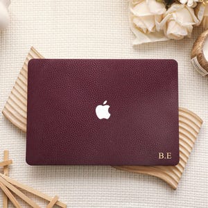 Puede incluir: Una funda para portátil de color burdeos oscuro con un acabado texturizado similar al cuero granulado. La funda presenta un recorte central para el logotipo de Apple y las iniciales doradas B.E. impresas en la esquina inferior derecha.