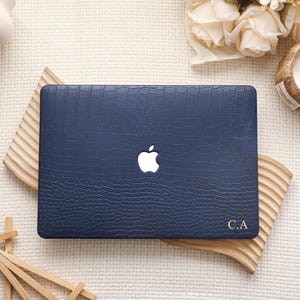 Può includere: Una custodia rigida per laptop blu navy scuro con un motivo testurizzato in pelle di coccodrillo. La custodia presenta un ritaglio centrale per il logo Apple bianco e le iniziali dorate C.A. stampate nell'angolo in basso a destra.