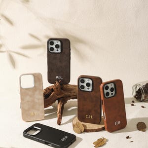 Może przedstawiać: Kolekcja spersonalizowanych etui na telefony w różnych kolorach, w tym brązowym, czarnym i beżowym. Każde etui ma unikalne inicjały w kontrastowym kolorze. Etui są ułożone na neutralnym tle.