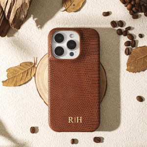 May include: Brown textured phone case with the initials "R|H" in gold. The case is surrounded by coffee beans, leaves, and a piece of wood, creating a natural aesthetic. The case appears to be made of leather or a similar material.