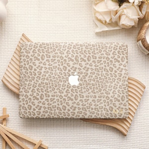 May include: A rectangular laptop hard shell case featuring a beige and brown leopard print pattern. The case has a central cutout for an Apple logo and the gold-colored initials M.P. printed in the bottom right corner of the cover surface.