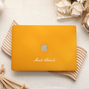 May include: A mustard yellow laptop hard shell case featuring a silver Apple logo in the center and the name Mark Aldrich printed in white script font below the logo. The case has a smooth matte finish and covers the top lid of the laptop.