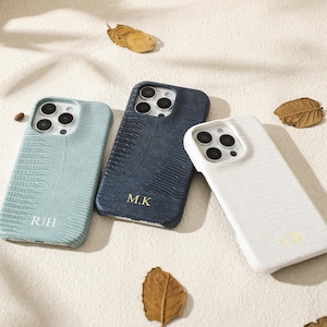 Puede incluir: Tres fundas de teléfono texturizadas en azul claro, azul marino y blanco, cada una con un monograma de inicial diferente. Las fundas están dispuestas sobre una superficie neutra con granos de café y hojas secas esparcidas.