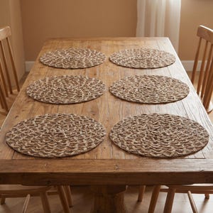 Puede incluir: Seis manteles individuales ovalados tejidos en color beige natural están colocados sobre una mesa de madera. Los manteles individuales tienen un patrón texturizado y circular. La mesa es de madera marrón claro y tiene un aspecto rústico. La escena sugiere un comedor o una cocina.