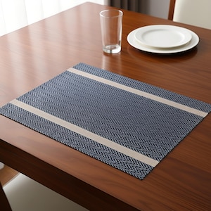 Puede incluir: Un mantel individual azul marino con un patrón de espiga y dos franjas beige. Un vaso transparente y dos platos blancos están sobre una mesa de madera. El mantel individual es rectangular.