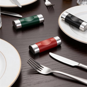 May include: A dining table setting with three cylindrical knife rests in green, red, and black. Each rest has a silver metal trim. White plates and silverware are arranged on a dark wooden table, creating a stylish look.