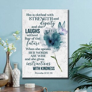 May include: A rectangular canvas print with inspirational text and floral art. The text reads "She is clothed with STRENGTH and dignity..." in various fonts. A blue flower and butterfly are depicted on the right. The bottom right corner has the Bible verse Proverbs 31:25-26.