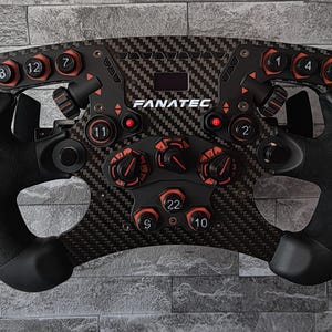 Könnte beinhalten: Ein schwarzes und Karbonfaser-Rennlenkrad mit mehreren roten Knöpfen, Drehreglern und einem kleinen Display. Das Lenkrad hat einen gepolsterten Griff und der Markenname "FANATEC" ist sichtbar. Der Hintergrund ist eine graue Backsteinmauer.