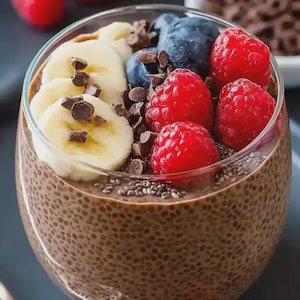 May include: A glass of chia seed pudding topped with fresh raspberries, blueberries, banana slices, and chocolate chips. The pudding is a rich brown color, and the fruit adds vibrant red and blue hues. A healthy and colorful dessert.