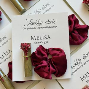 May include: A display featuring deep red scrunchies, small glass vials containing green powder and dried flowers, and white cards with the text "Teşekkür ederiz" and "MELISA Henna Night".