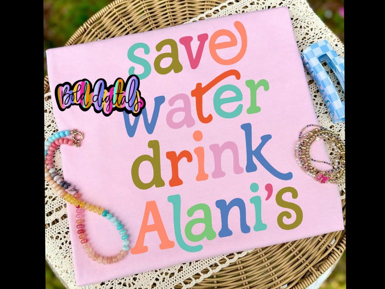 Puede incluir: Una camiseta rosa claro con las palabras "save water drink Alani's" en varios colores. La camiseta se muestra sobre una superficie de mimbre con un collar de cuentas y otros accesorios.