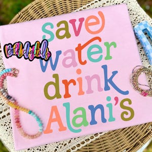 Puede incluir: Una camiseta rosa claro con las palabras "save water drink Alani's" en varios colores. La camiseta se muestra sobre una superficie de mimbre con un collar de cuentas y otros accesorios.