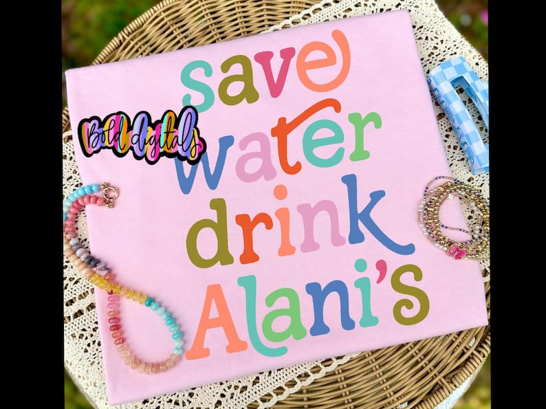 Puede incluir: Una camiseta rosa claro con las palabras "save water drink Alani's" en varios colores. La camiseta se muestra sobre una superficie de mimbre con un collar de cuentas y otros accesorios.
