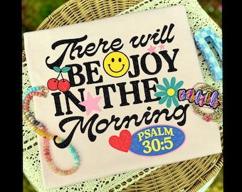 May include: A light beige t-shirt with the phrase "There will be JOY IN THE MORNING" in black script. The design includes a yellow smiley face, cherries, a pink star, a blue flower, and a red heart. A colorful beaded necklace and a gold bracelet are also visible.